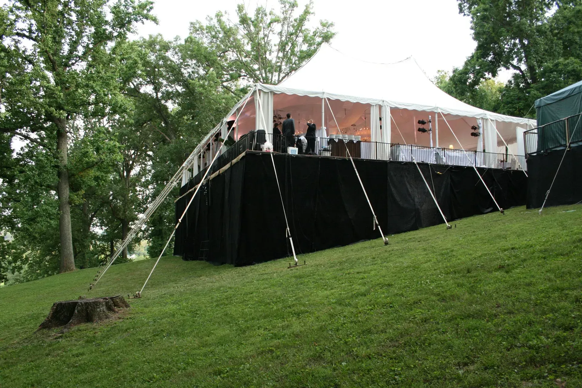 A large white event tent elevated on a platform with black skirting, set on a grassy hill surrounded by trees.