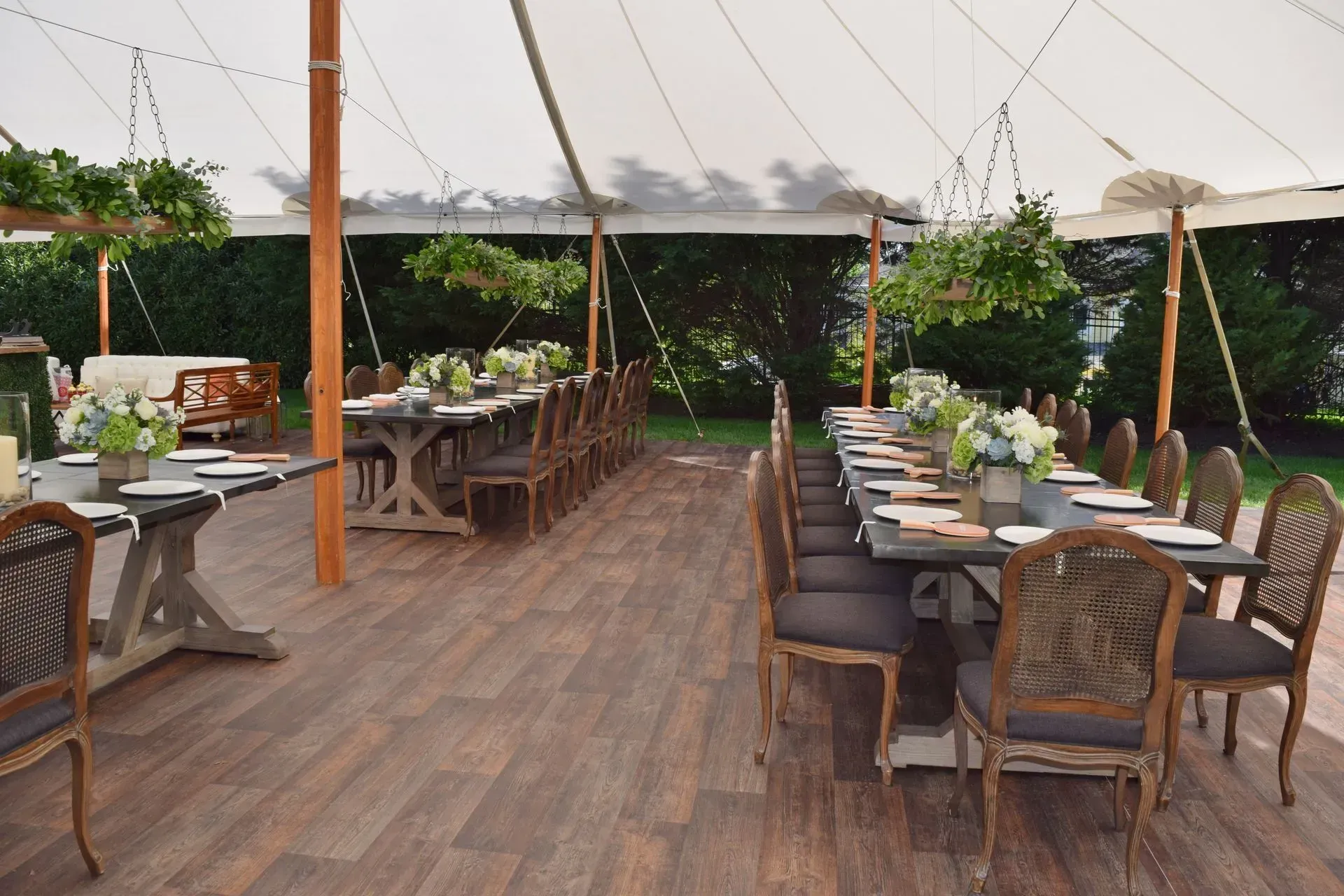 Tables set for a formal event under a large white tent on a wood-look floor, with hanging greenery and wooden chairs.