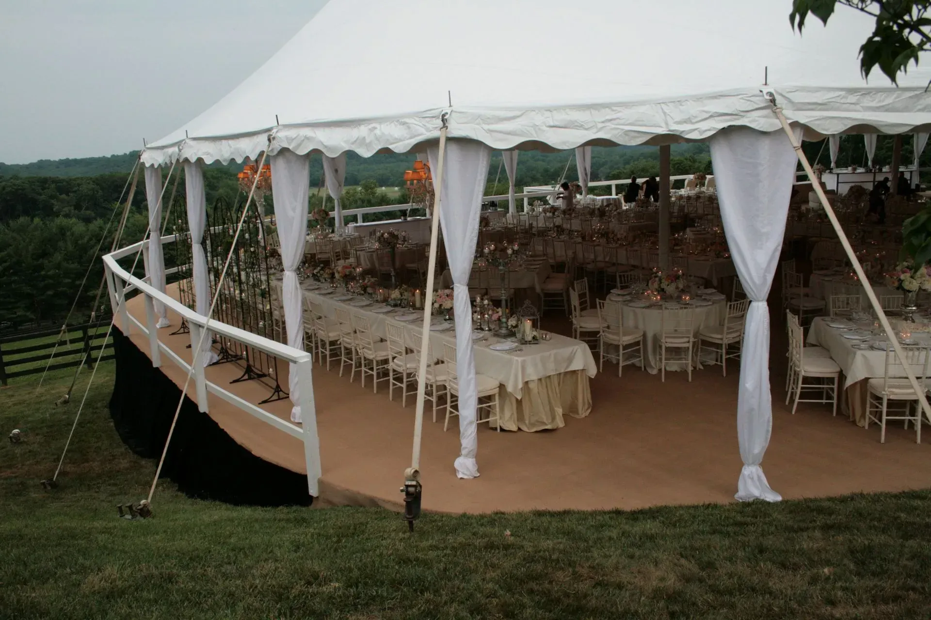 A large, white wedding tent set up outdoors on a hill, featuring draped fabric columns, tables, and views of a forest.