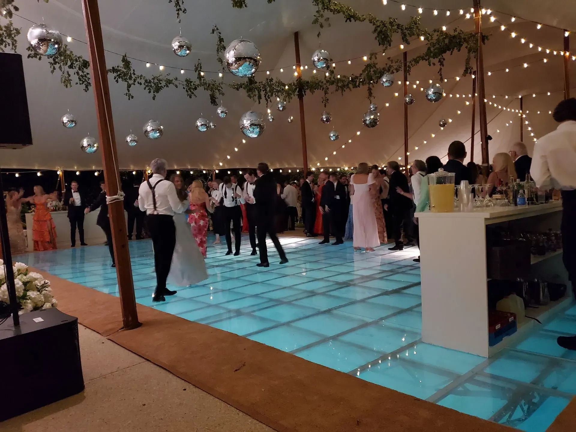 Guests dance on an illuminated blue floor in a tent decorated with hanging disco balls and strings of warm fairy lights.