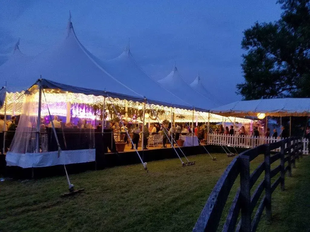 A large white tent with glowing string lights hosts an evening event, set against a dark blue sky and a wooden fence.