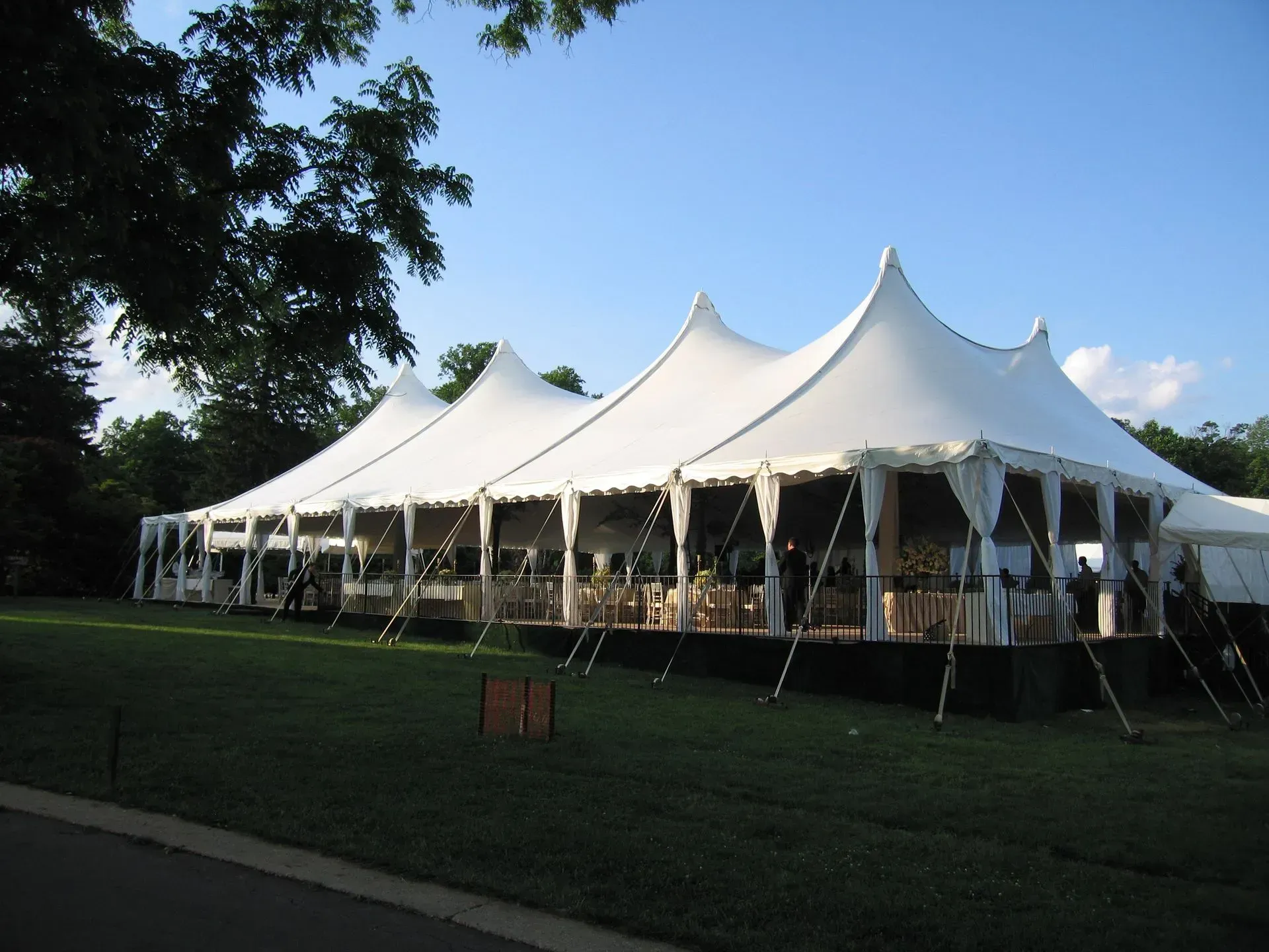 A large white high-peak event tent set up on a grassy lawn with trees in the background under a blue sky.