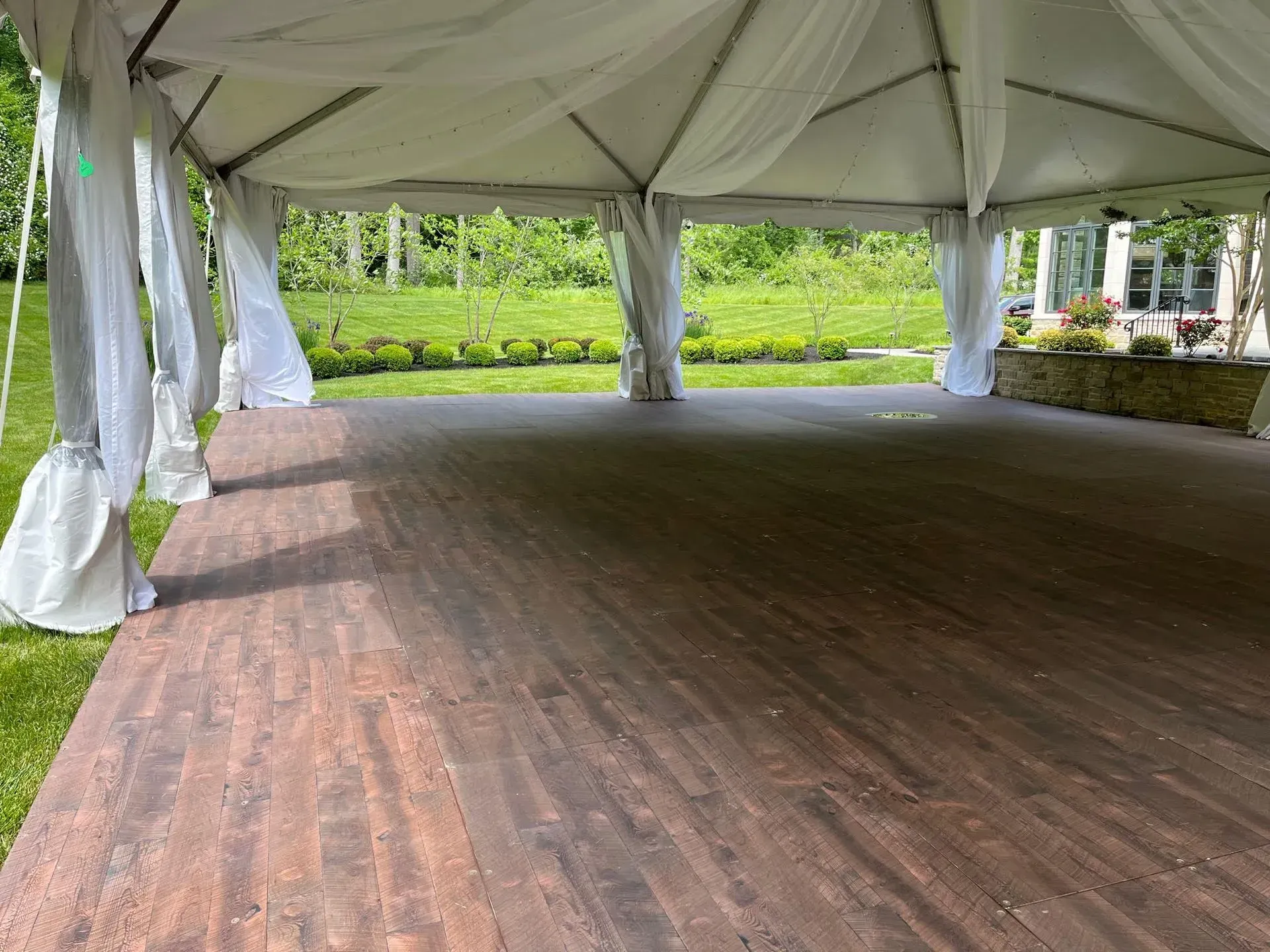 A large, white event tent with white fabric draping over a dark wood-look floor, set against a green outdoor landscape.
