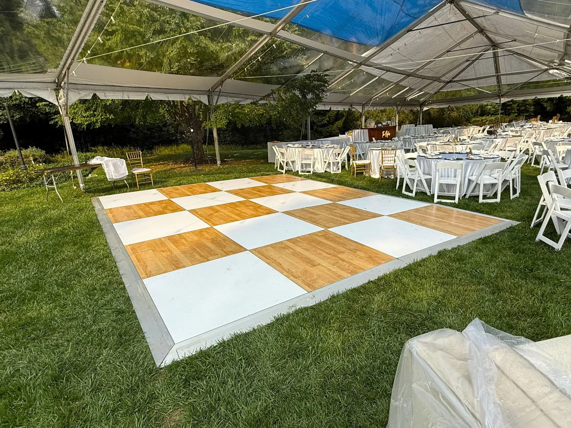White-and-wood dance floor under a tent, surrounded by white chairs on a grassy outdoor event lawn.