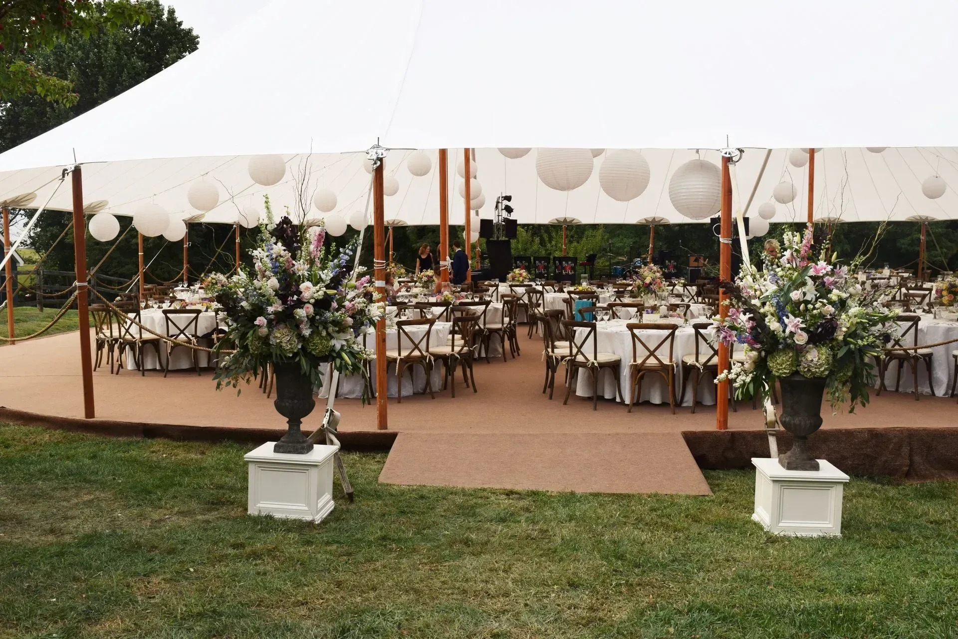 An outdoor wedding tent set up with round tables, white chairs, floral centerpieces, and hanging paper lanterns.