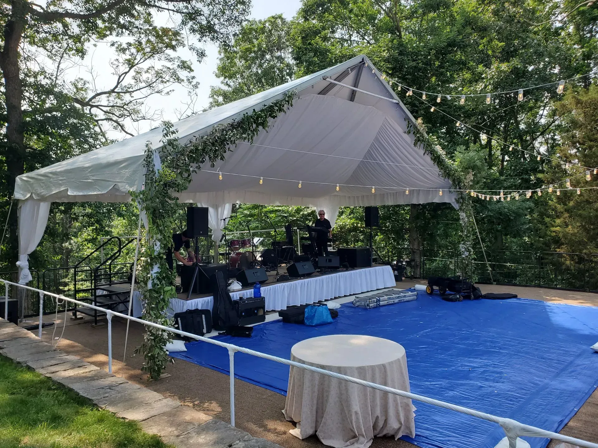 An outdoor event space features a white peaked tent, a blue dance floor, and a stage setup under a canopy of trees.