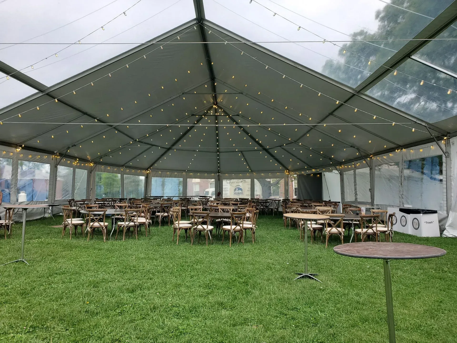 An outdoor event tent with string lights, set up with numerous round tables and chairs on a grassy field.