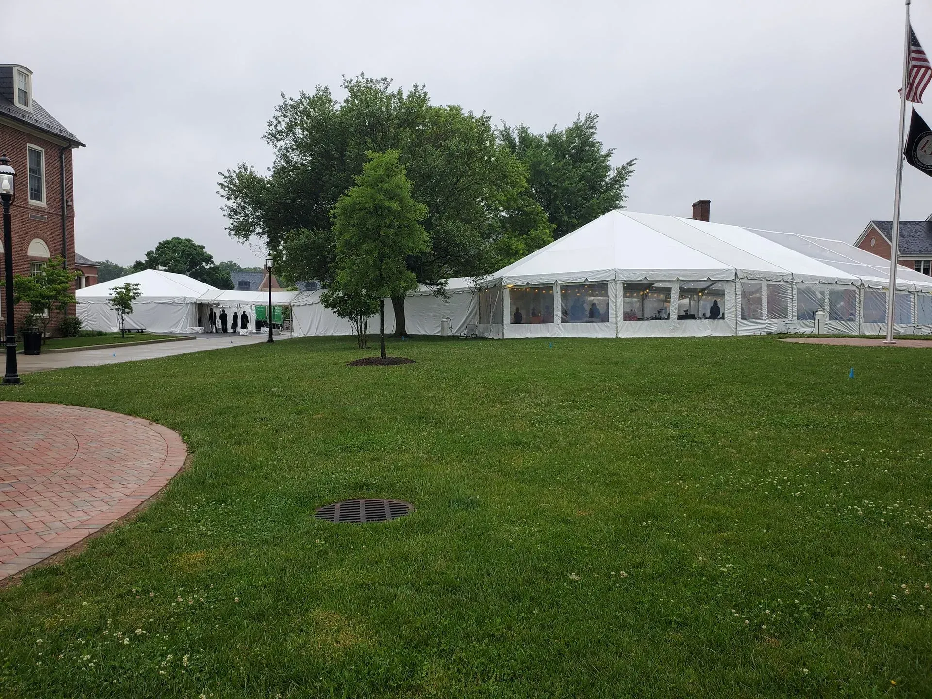 A large white event tent sits on a grassy lawn next to a brick building under a cloudy sky.