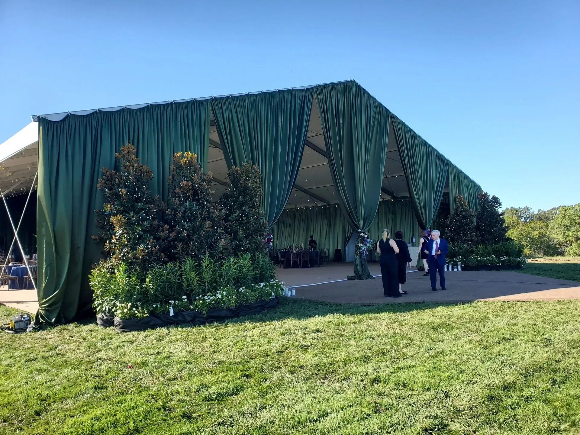 A large, open-air tent with dark green curtains stands on a grassy field, with a few people standing near the entrance.