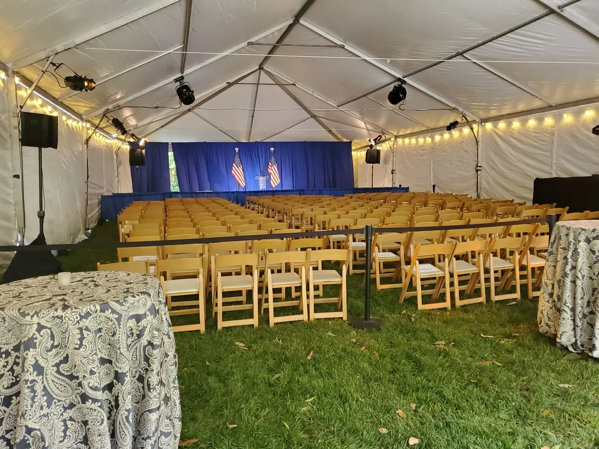Rows of wooden chairs arranged in a large, white-tented event space with a blue-curtained stage.