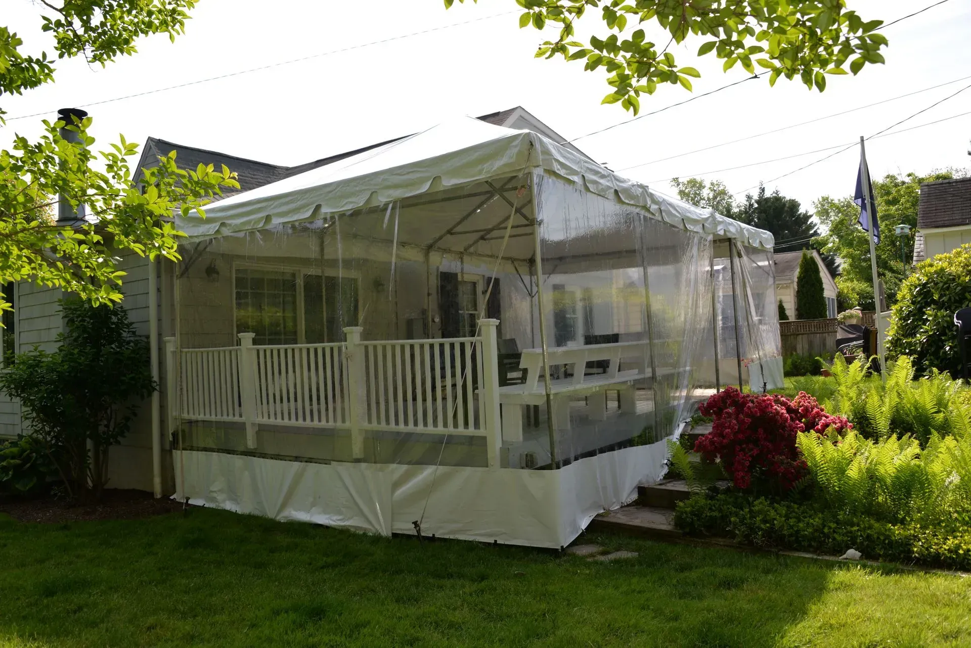 A white tent covering a residential deck, featuring clear plastic side panels, set against a green lawn and trees.