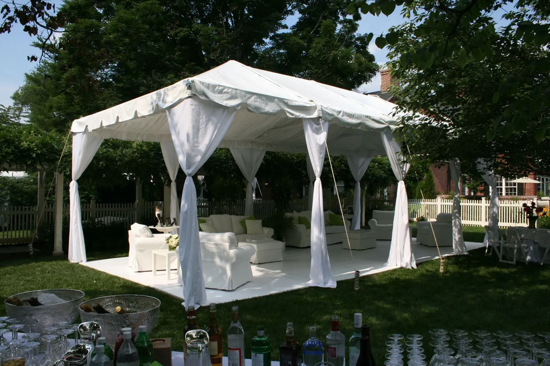 White canopy tent with draped fabric pillars over outdoor lounge furniture on a green lawn, with a bar setup in front.