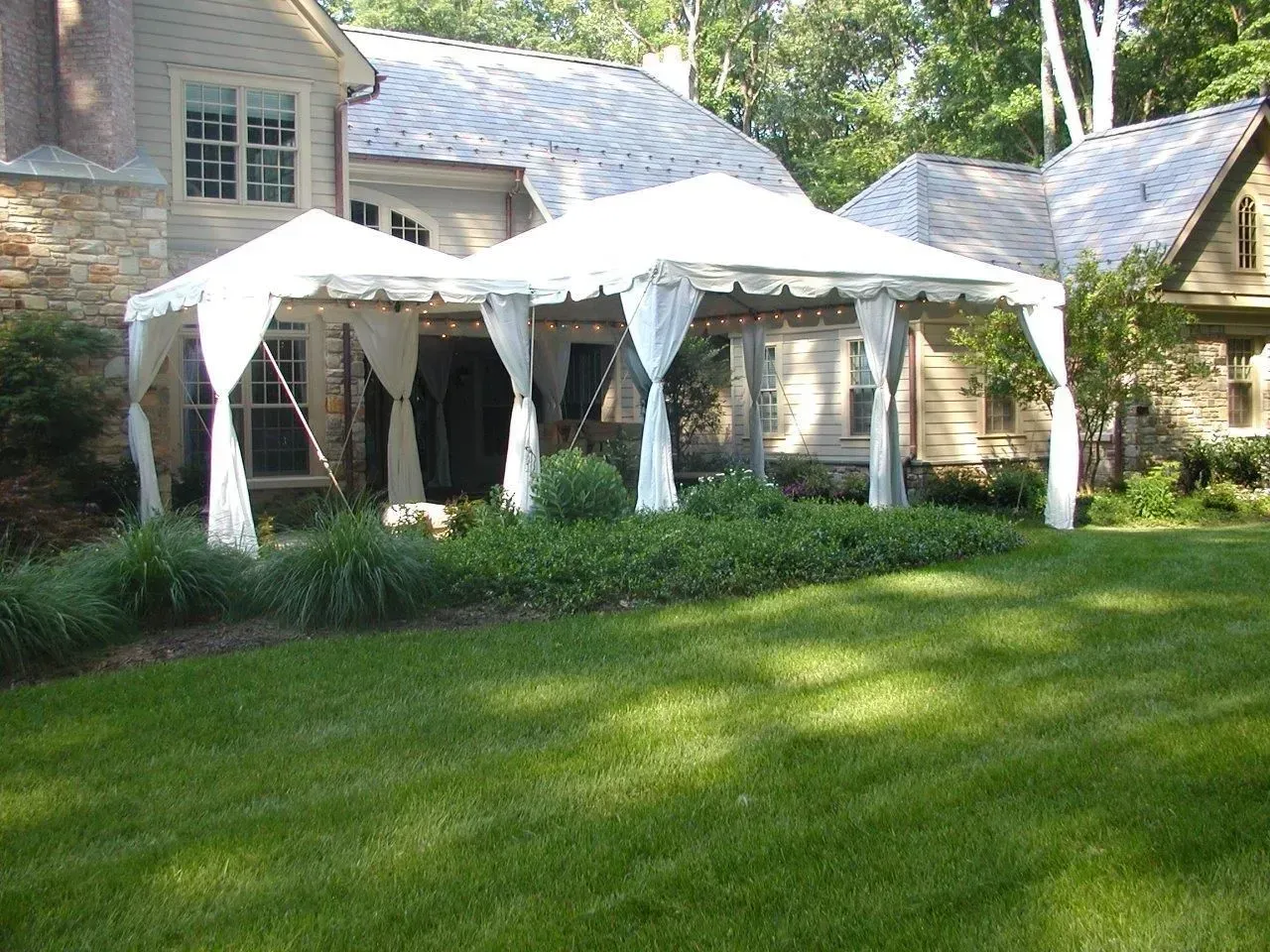 Two white wedding-style tents set up on a lush green lawn in front of a stone house with decorative fabric drapes.