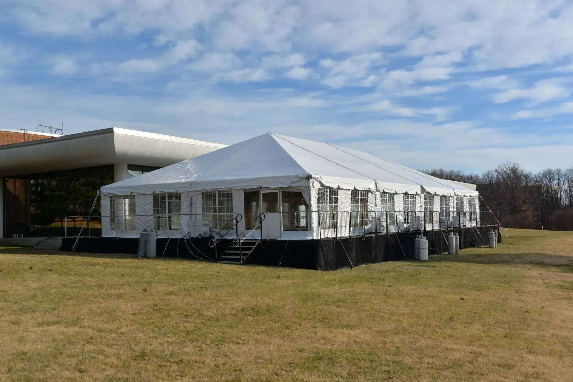 A large, white event tent with clear window panels sits on a black platform in a grassy field next to a building.