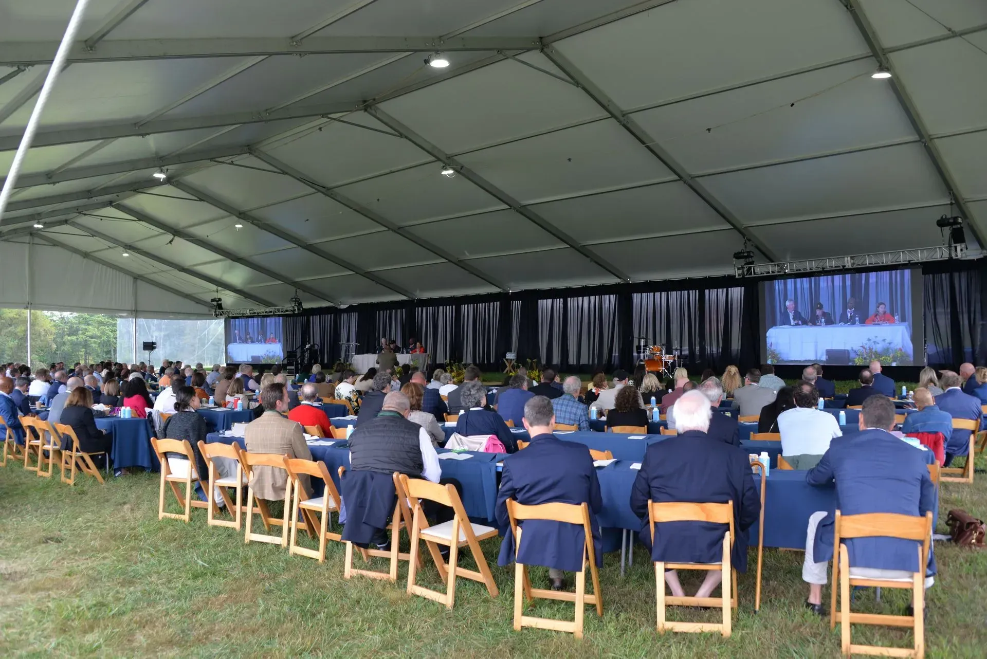 A large crowd sits at tables under a tent during an outdoor event with a presentation screen at the front.