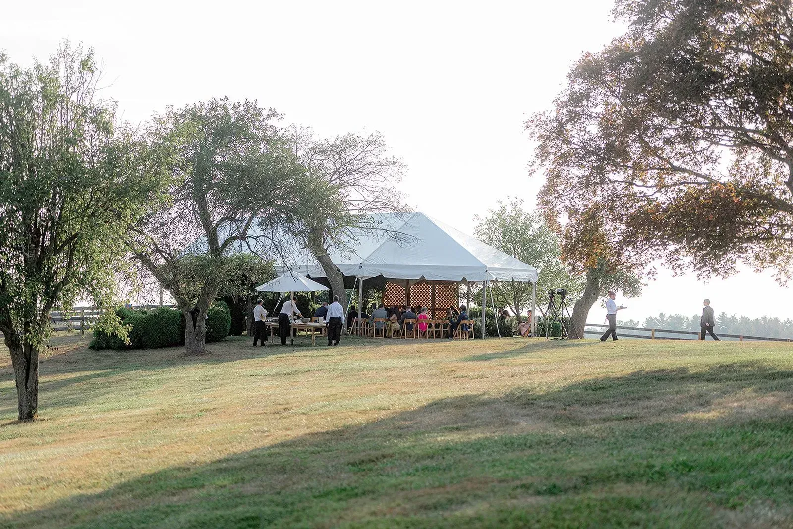 A white tent hosts a gathering on a grassy hill, surrounded by trees, with people walking nearby on a sunny day.