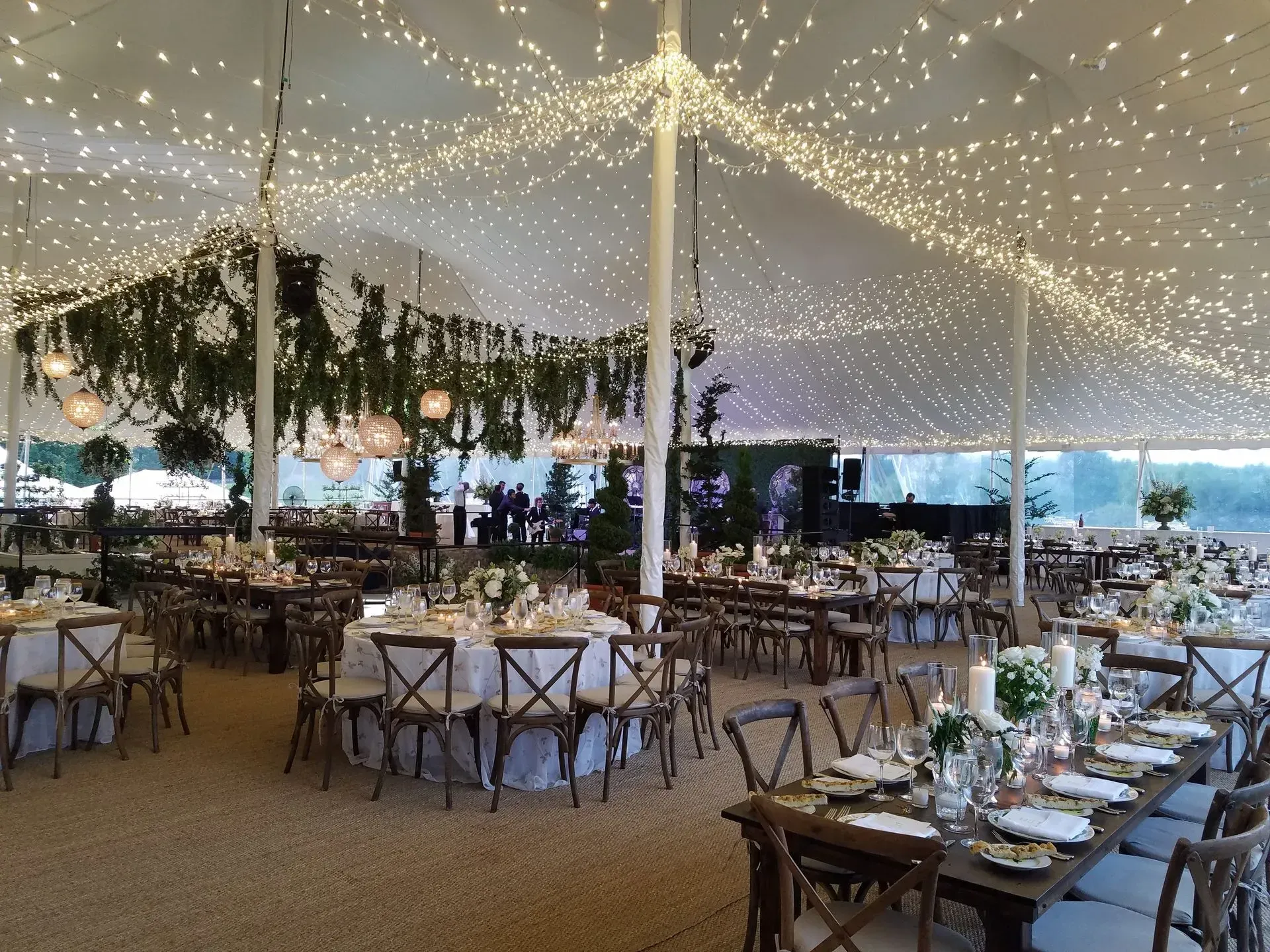 A tented event space featuring tables with chairs and a ceiling covered in strings of glowing fairy lights and greenery.