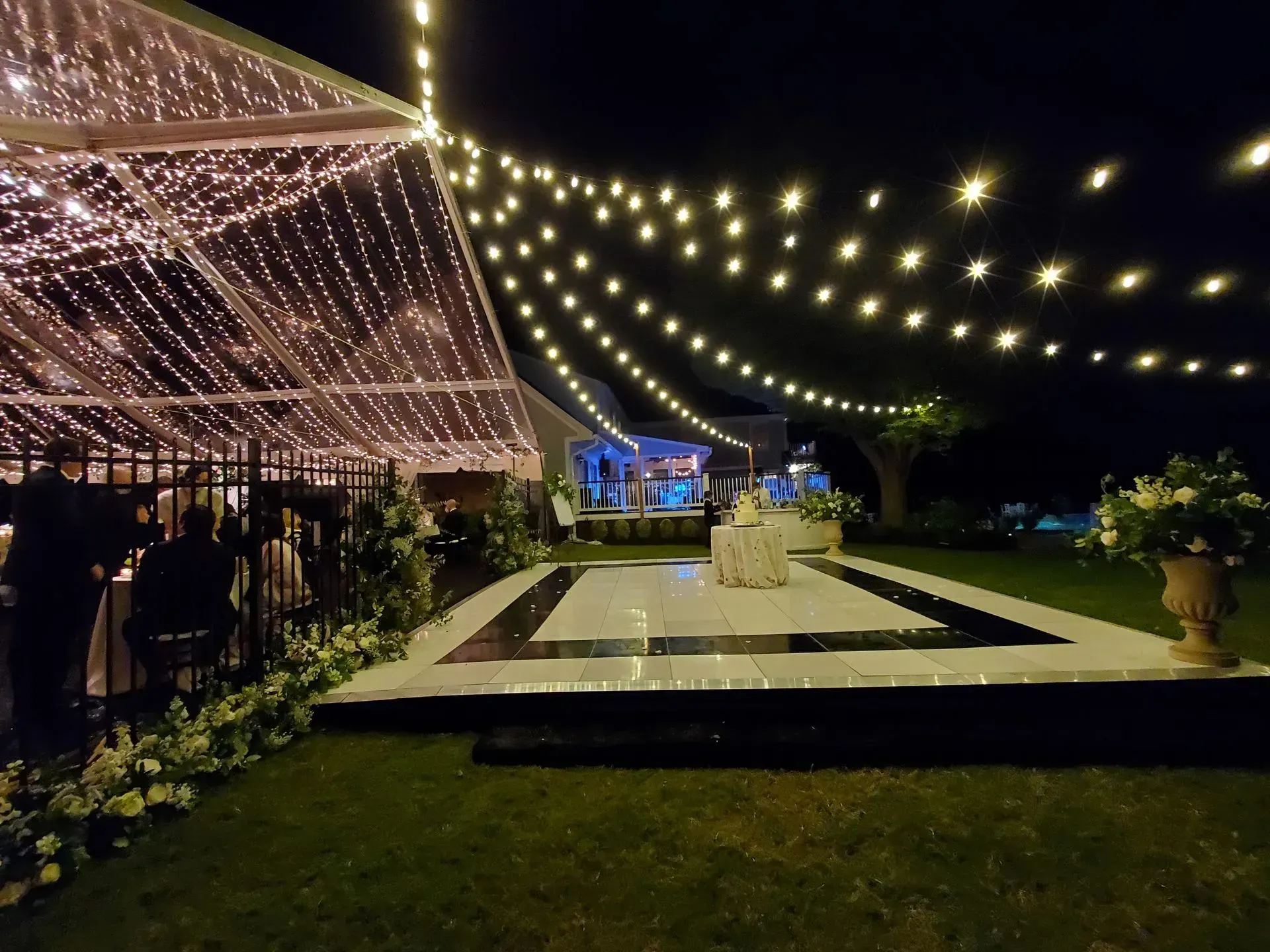 An outdoor night event with a lit dance floor, white tent, and strings of fairy lights overhead on a grass lawn.
