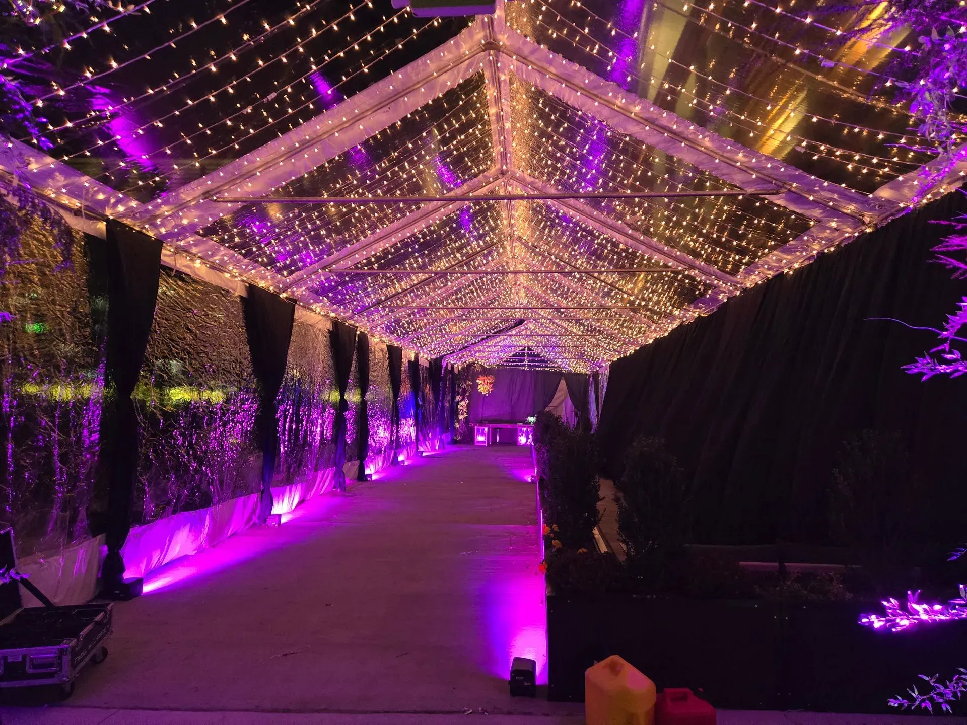 An outdoor tent at night, decorated with strings of fairy lights, purple accent lighting, and black fabric drapes.