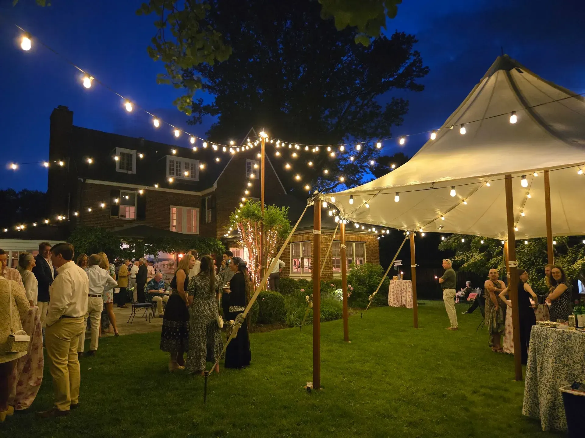 People mingle on a lawn at dusk under strings of warm lights and a large white canopy tent, with a brick home behind.