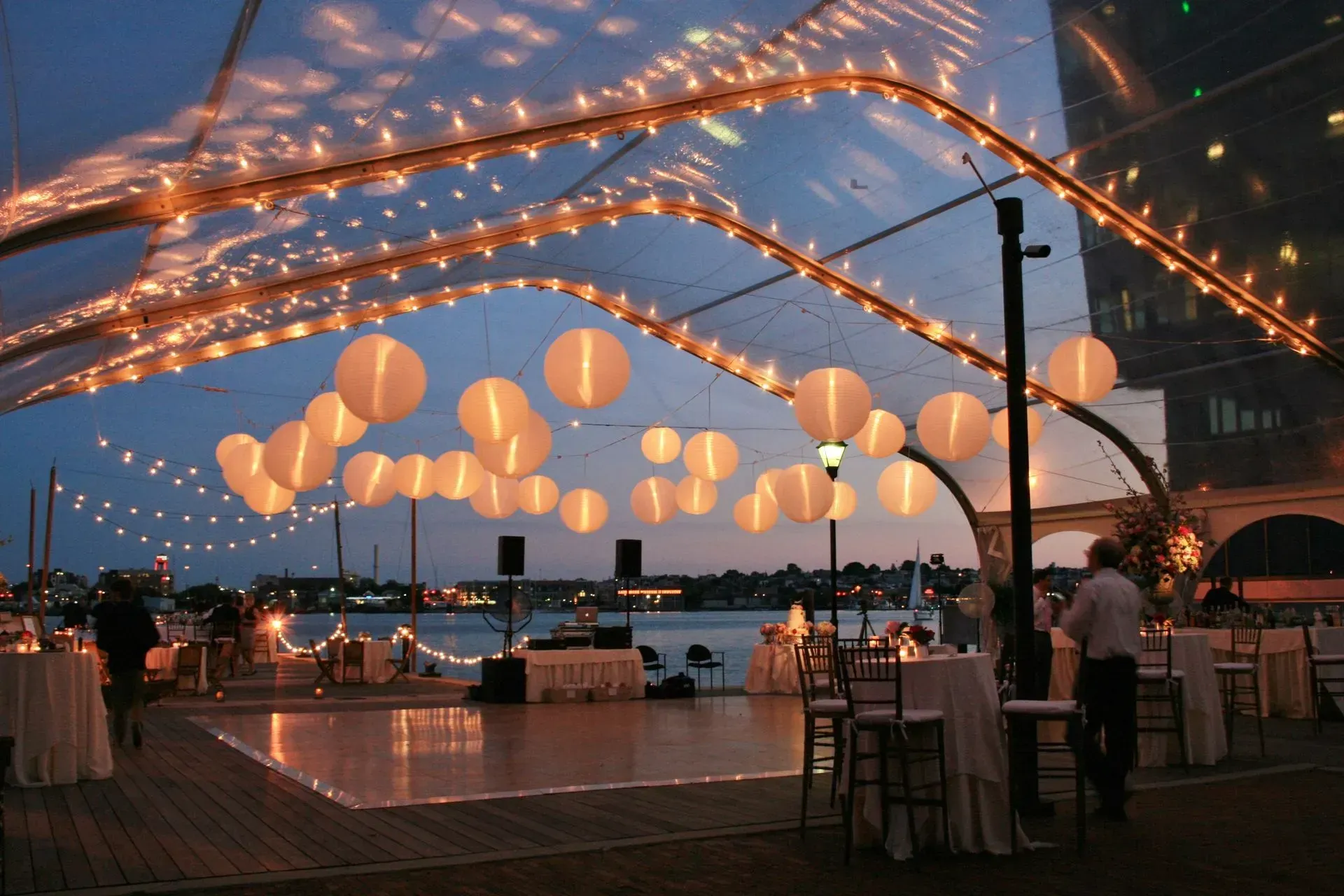 Outdoor evening event with a dance floor, white paper lanterns hanging from a tent frame, and string lights by water.