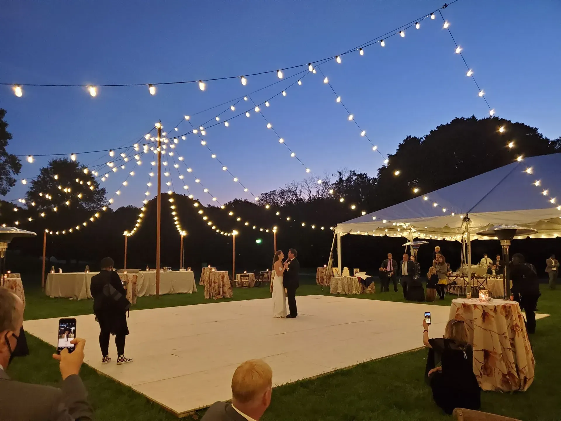 A couple dances on an outdoor white floor beneath string lights at dusk, surrounded by tables and guests.