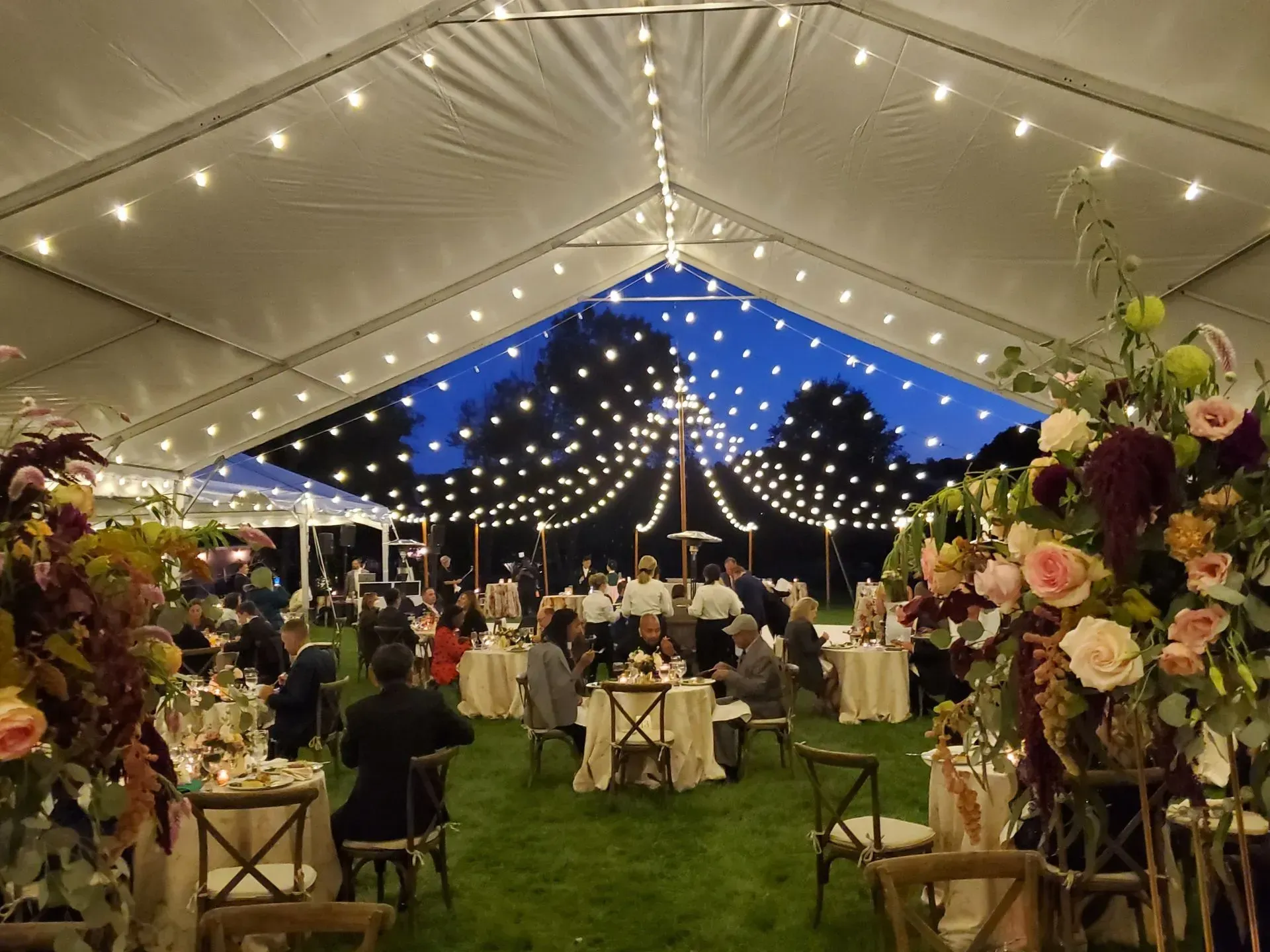 Outdoor event under a lit tent with tables, chairs, and large floral arrangements at dusk.