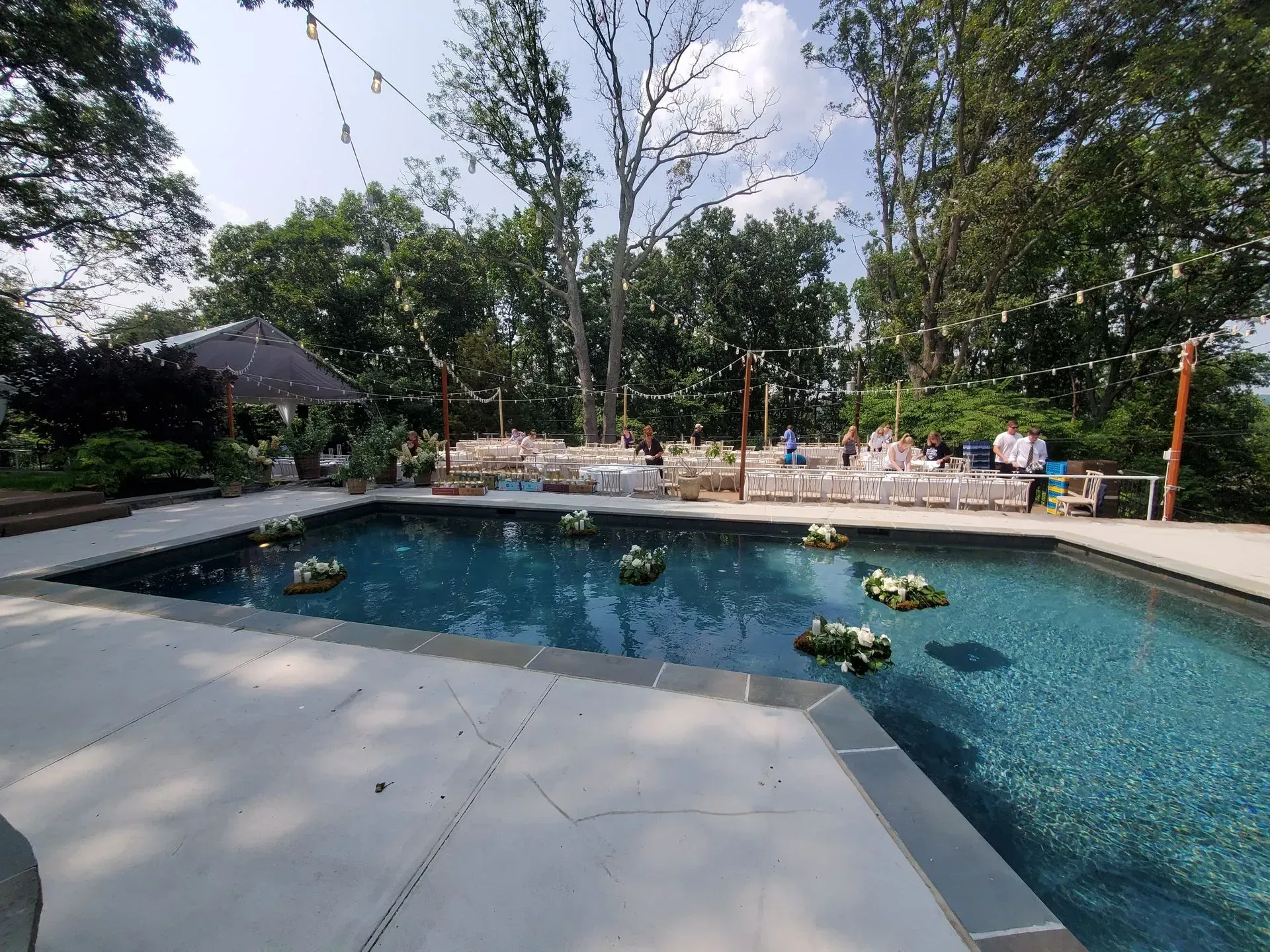 A backyard swimming pool with floating floral arrangements, surrounded by an outdoor event setup with tables and trees.