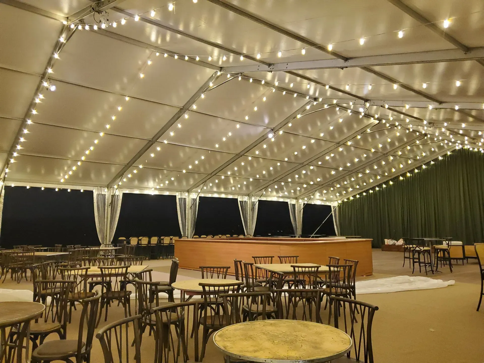 A large tent at night featuring round tables, wooden chairs, string lights on the ceiling, and a wooden bar in the back.