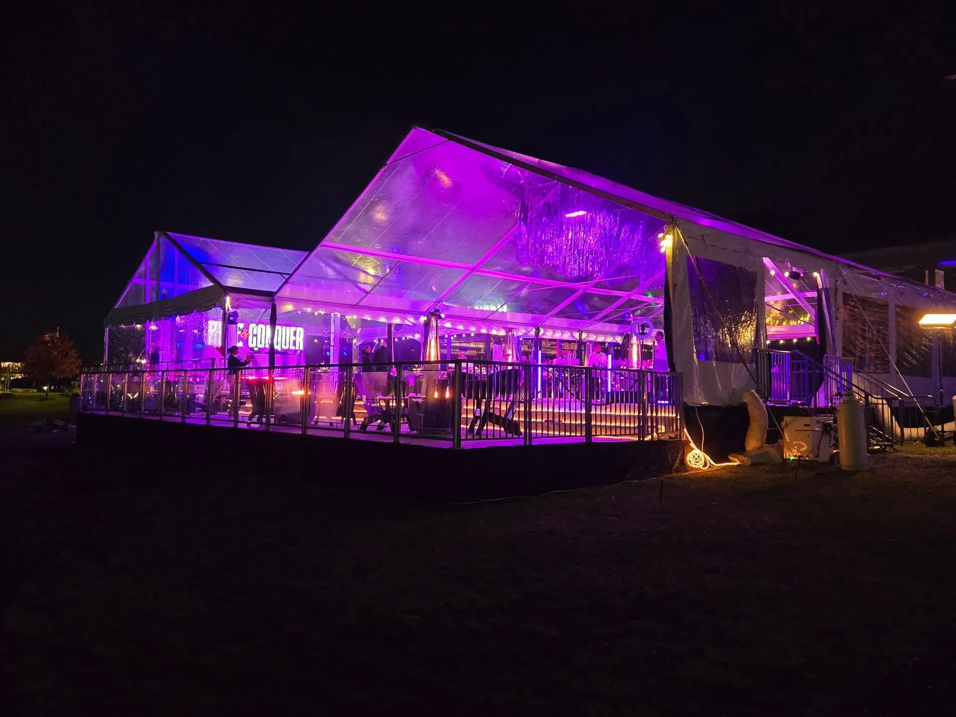 A large, clear-topped outdoor event tent illuminated with vibrant purple lighting at night.