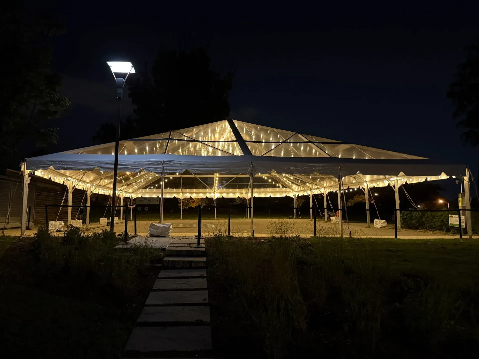 A large, illuminated event tent at night, featuring a path of stone pavers leading to the entrance in a grassy park setting.