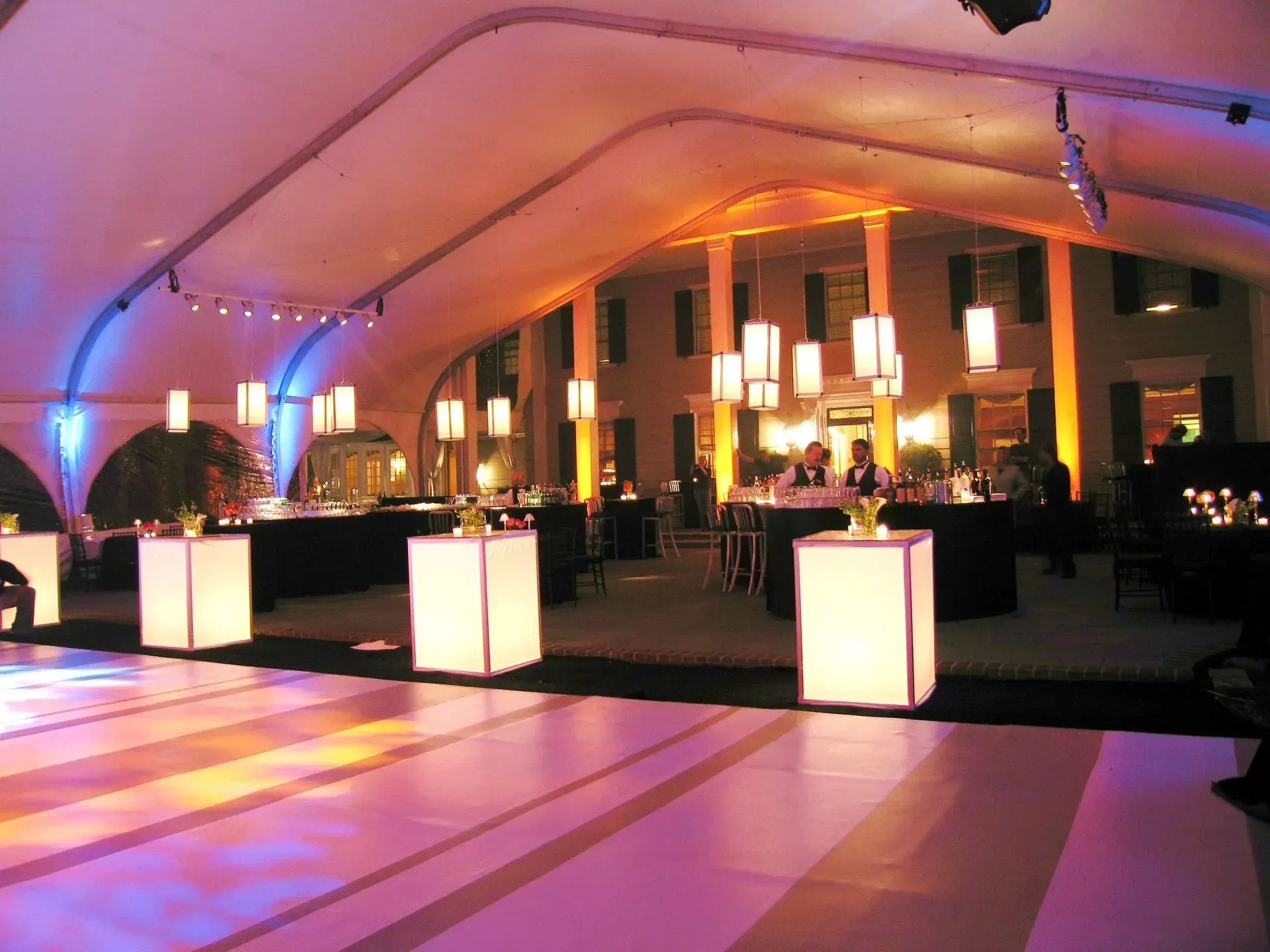 An event tent at night, featuring a dance floor, glowing cube tables, and ambient lighting in front of a building.