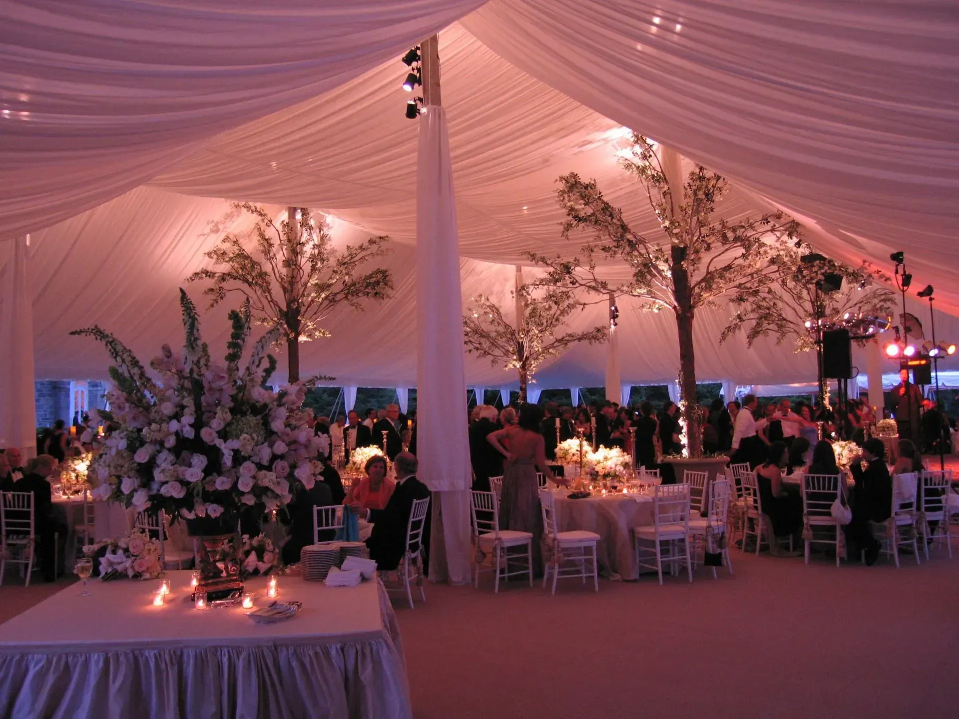An elegant event held under a large white tent with pink lighting, decorated with floral centerpieces and indoor trees.