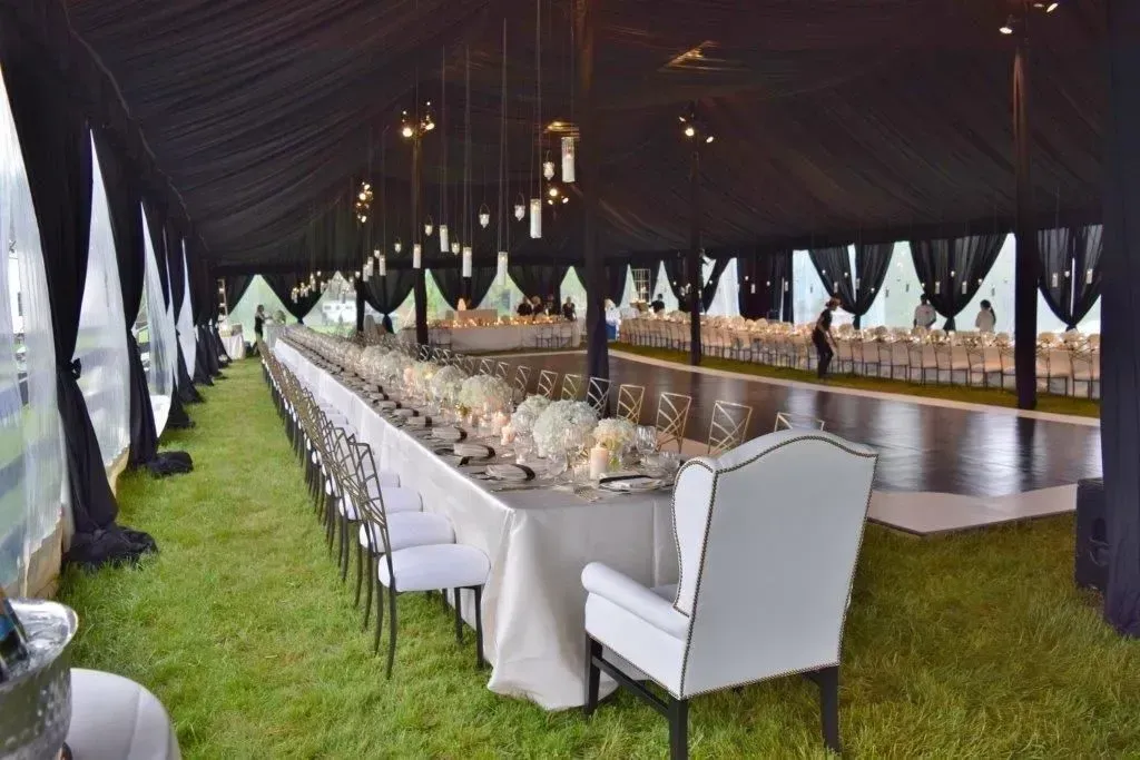 Long white tables set for a banquet inside a large, black-draped tent with a wooden dance floor and green grass floor.