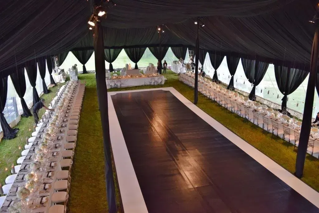 An elegant marquee setup with long banquet tables flanking a central dark dance floor under dark, draped tent ceilings.