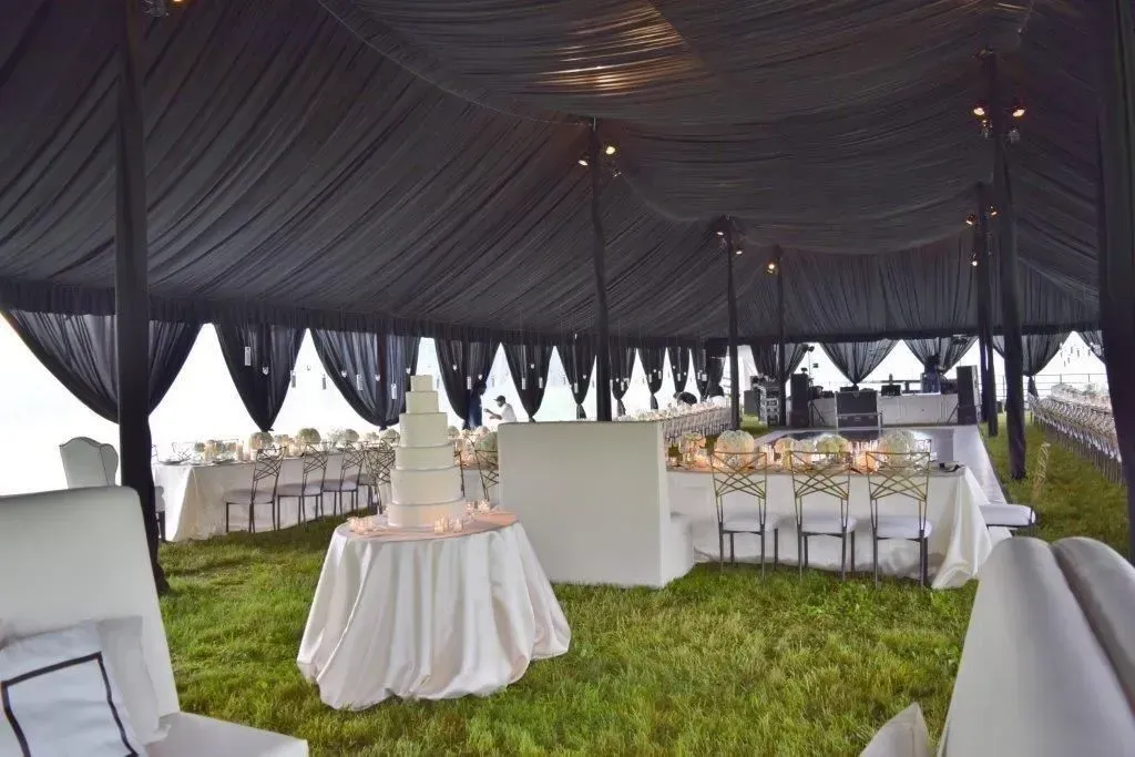 A white tiered wedding cake stands on a round table under a large tent with black draped ceiling fabric and lawn flooring.