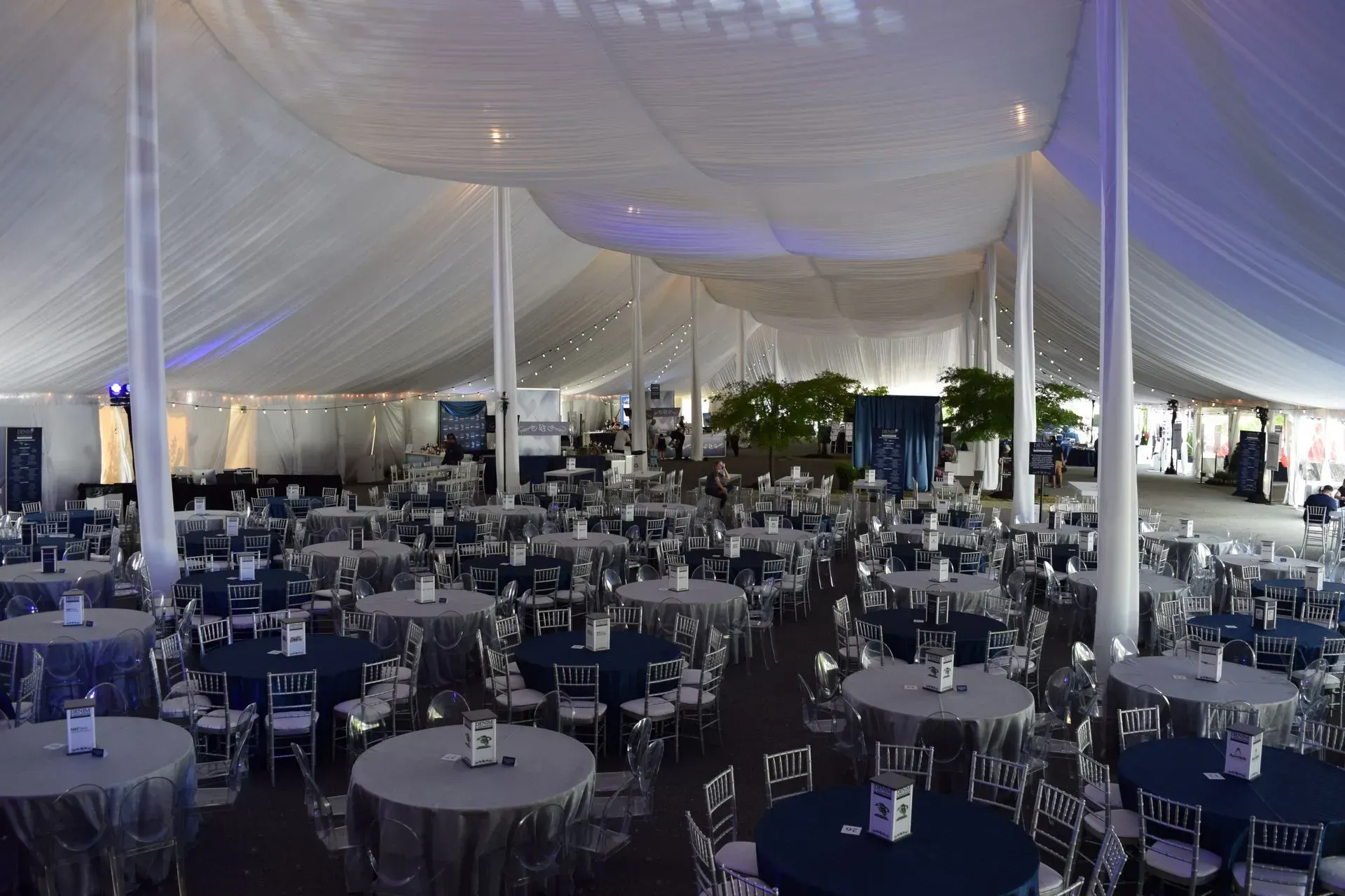 A large event tent filled with rows of round tables with navy blue and silver tablecloths and silver chairs.