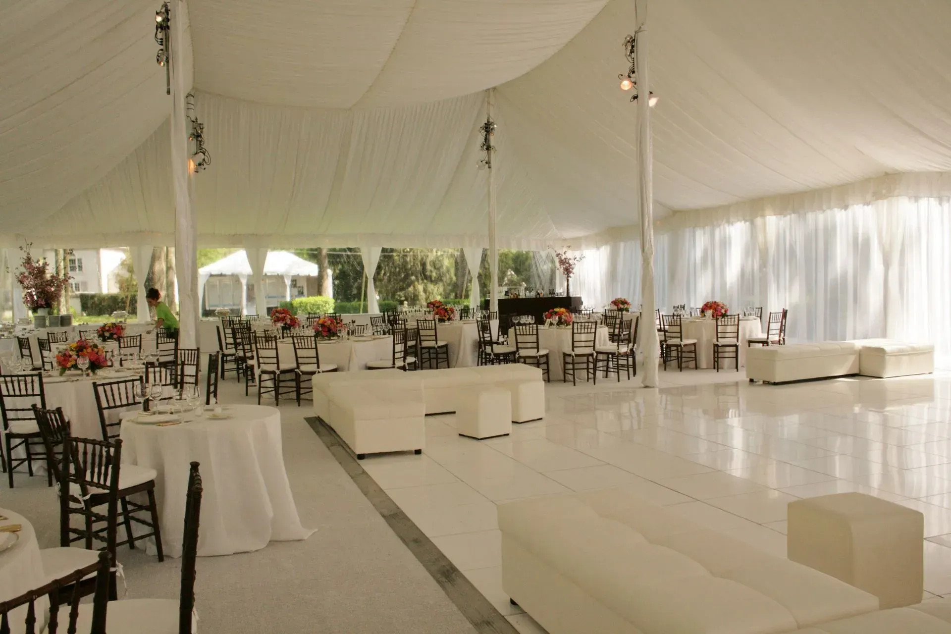 A brightly lit white event tent featuring round tables, chairs, and modern white lounge furniture on a glossy dance floor.