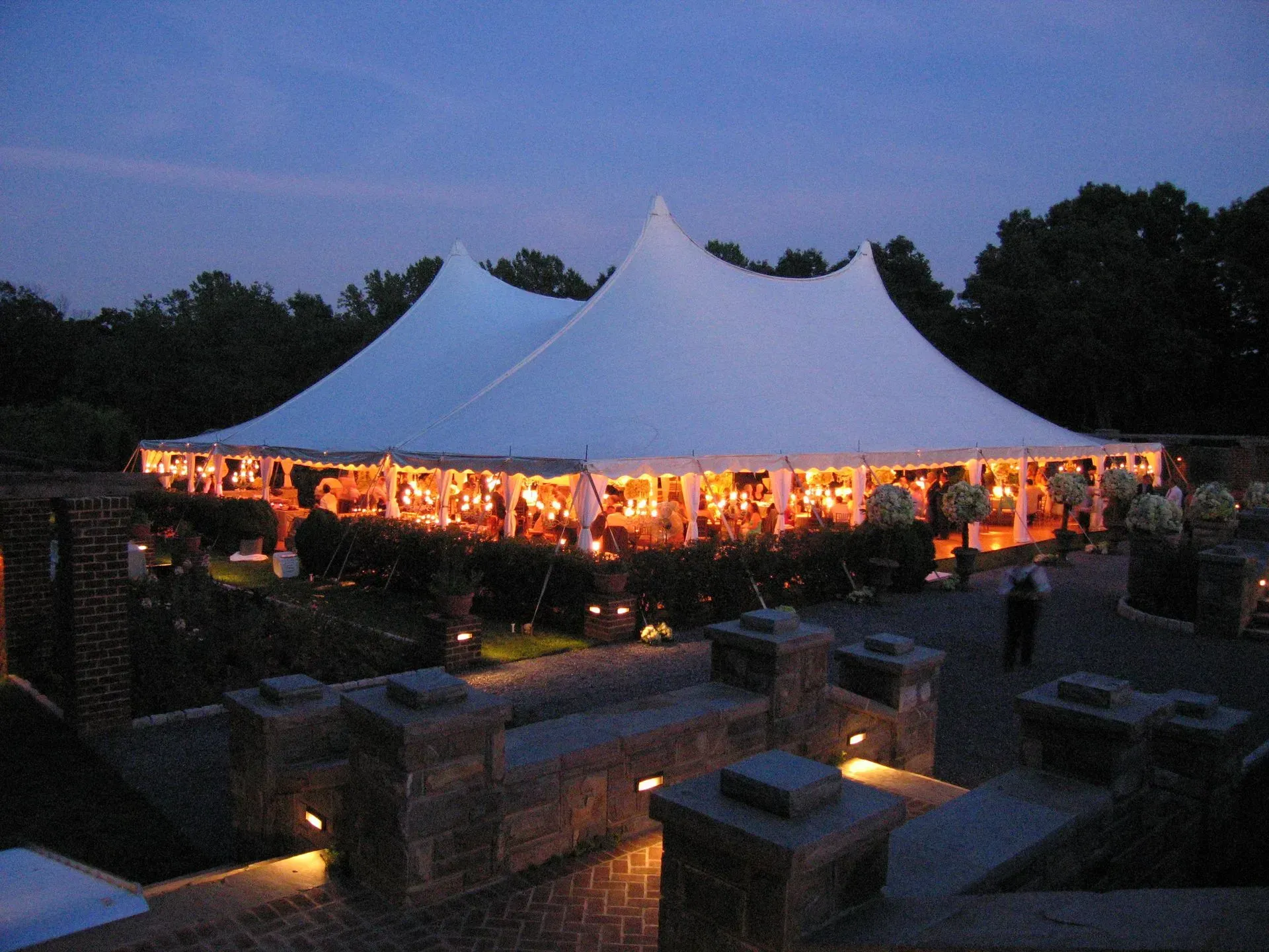 An illuminated white marquee tent glows at dusk, nestled in a dark, landscaped outdoor setting with stone pathways.