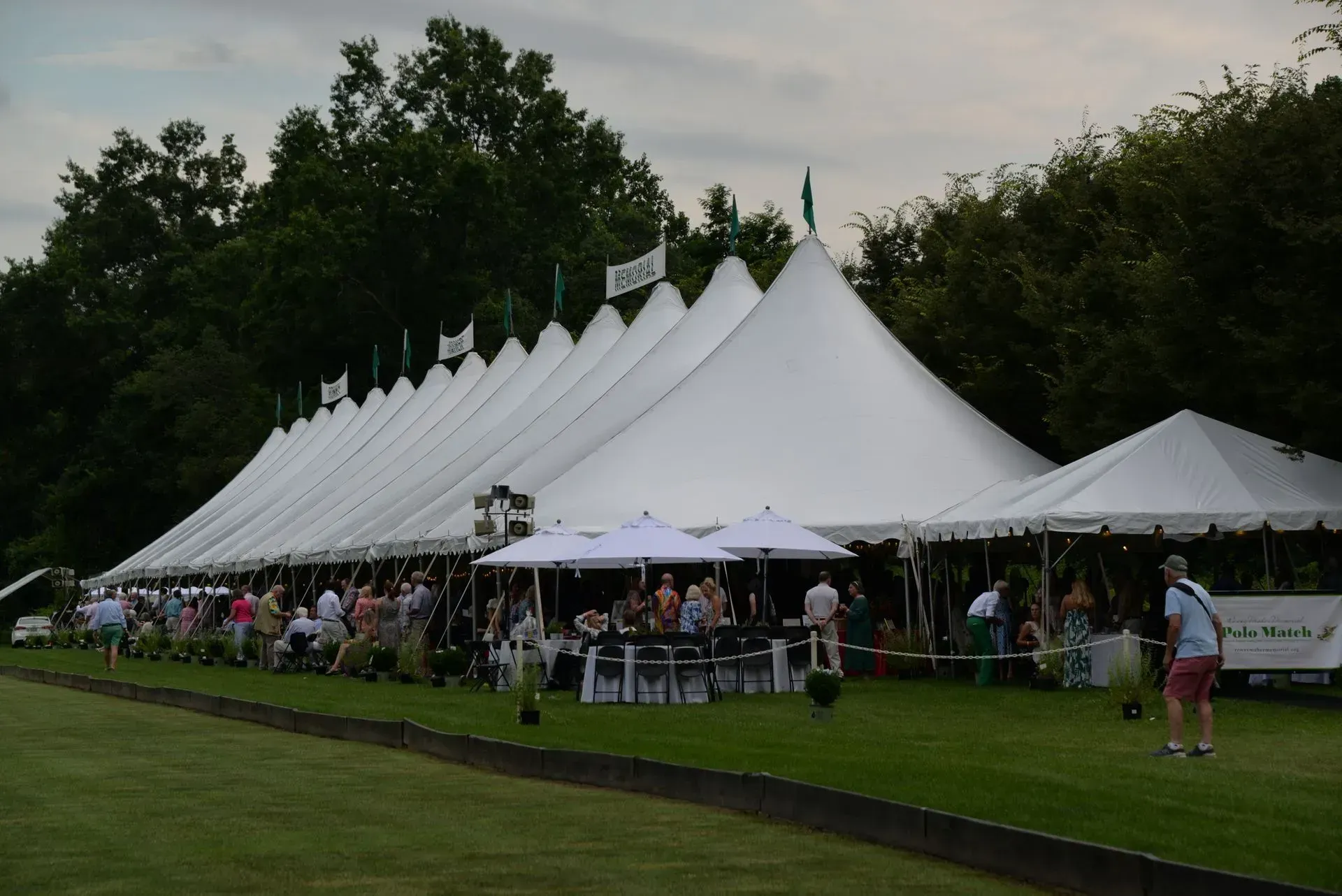 People gathered under a long row of connected white event tents on a grassy field at dusk.