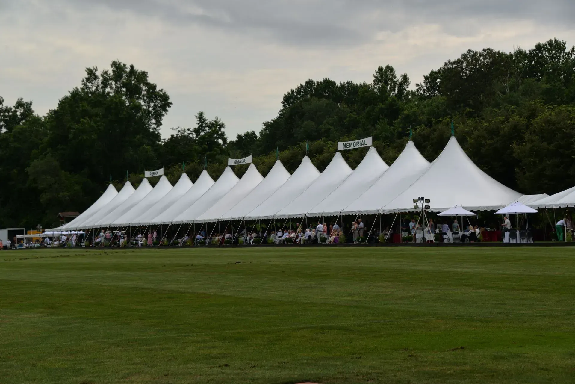 A long, white, peaked event tent set up on a grassy field with trees in the background.