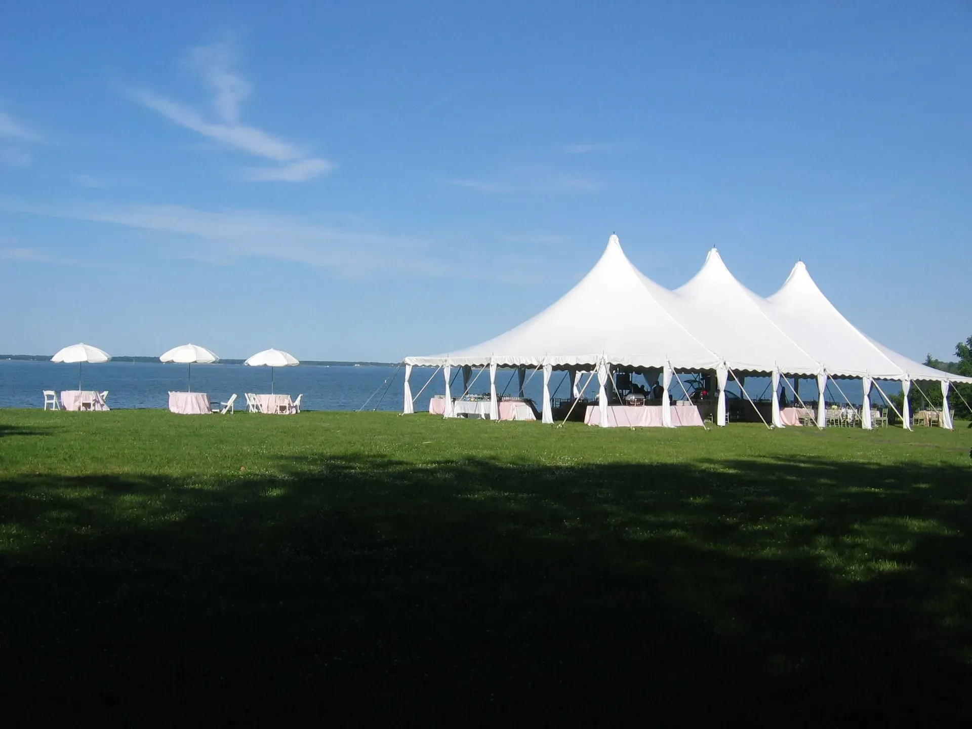 Large white event tent on a grassy lawn by a lake, with three smaller patio umbrellas set up along the water.