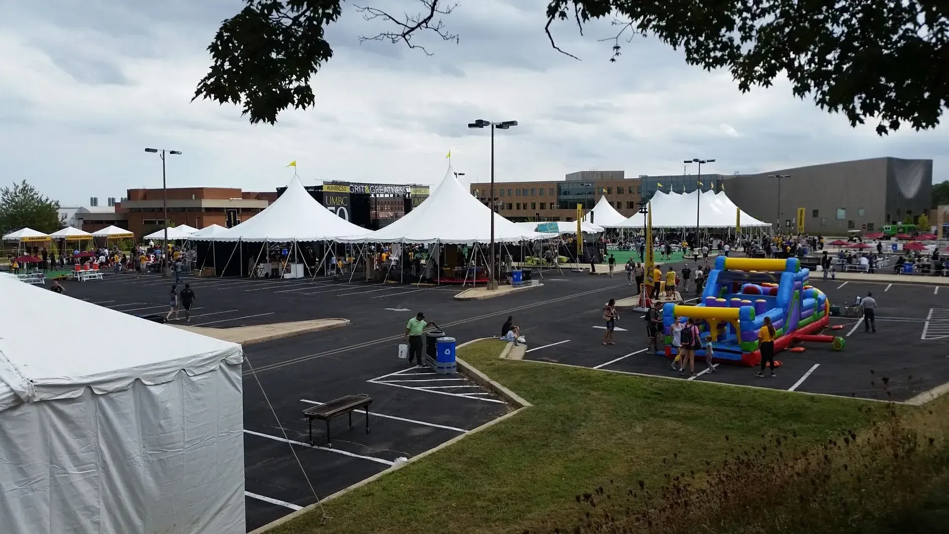 An outdoor community festival in a parking lot with white event tents, a bounce house, and people walking around.