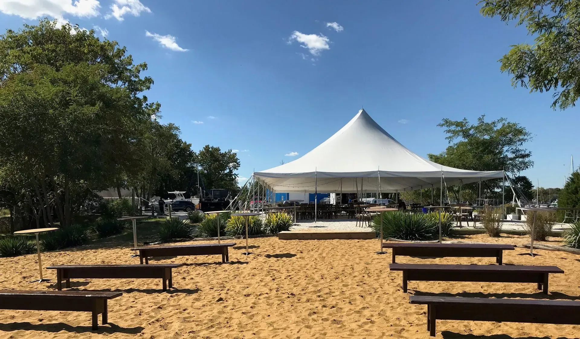 A large white event tent stands in a sandy clearing with wooden benches arranged in rows under a clear blue sky.