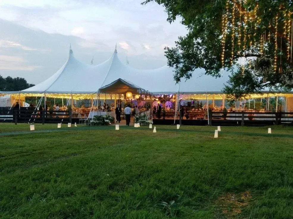 A large white event tent lit for an evening reception on a grassy lawn with glowing lanterns along a path.