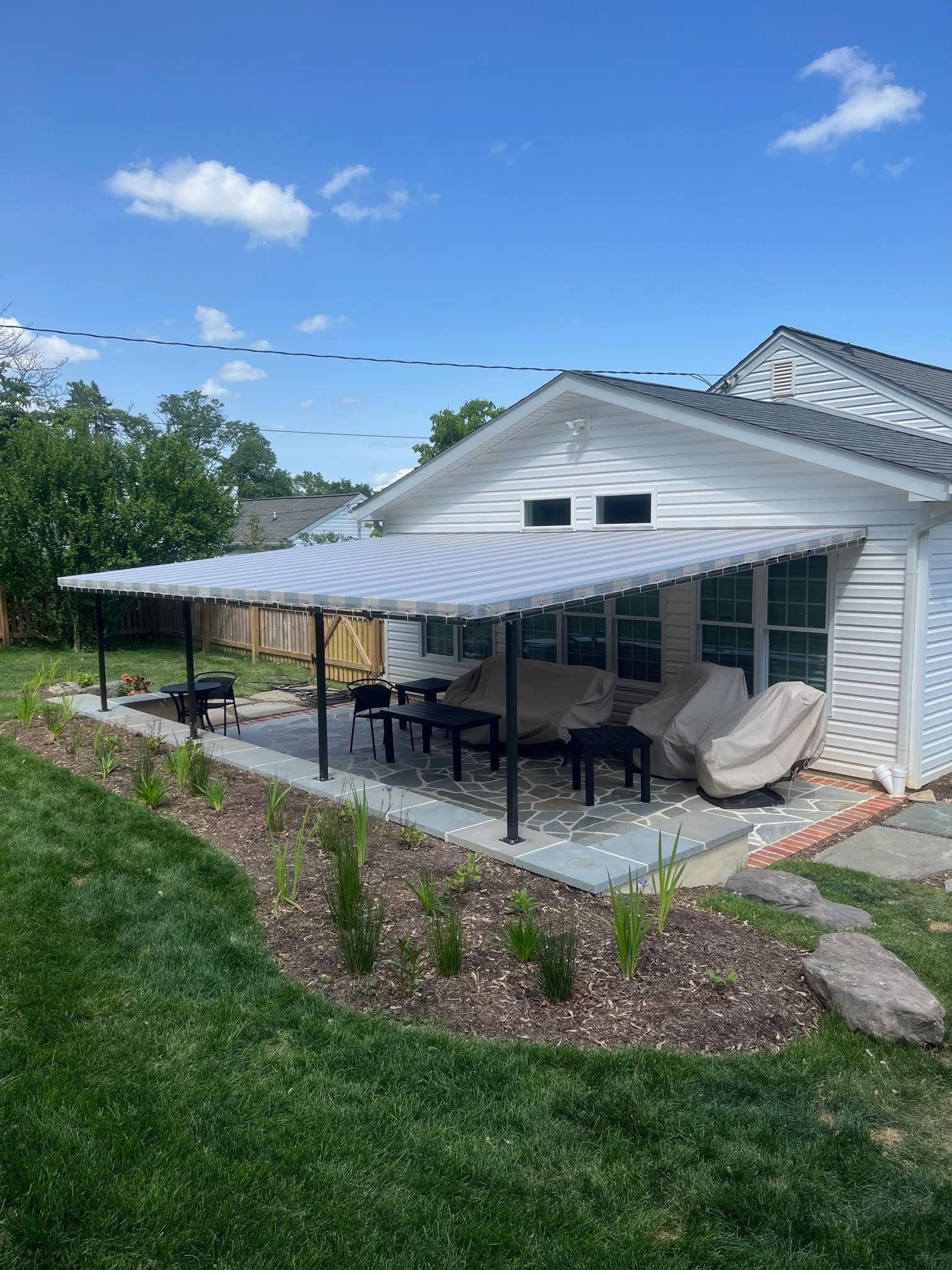 Backyard patio with black awning, outdoor dining table, chairs, and a hanging egg chair beside a white house