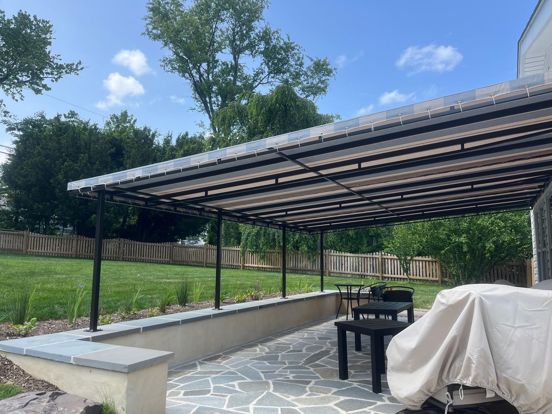 Covered backyard patio with stone flooring, black metal posts, and a grill under a striped awning.