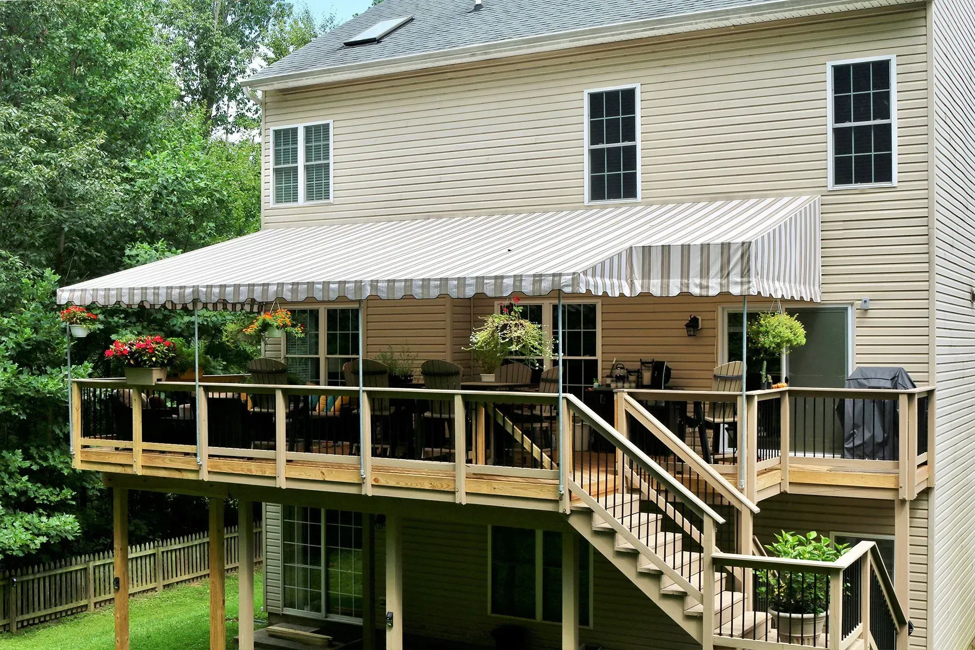 A beige two-story house with a wooden deck, stairs, and a white striped retractable awning in a lush backyard.