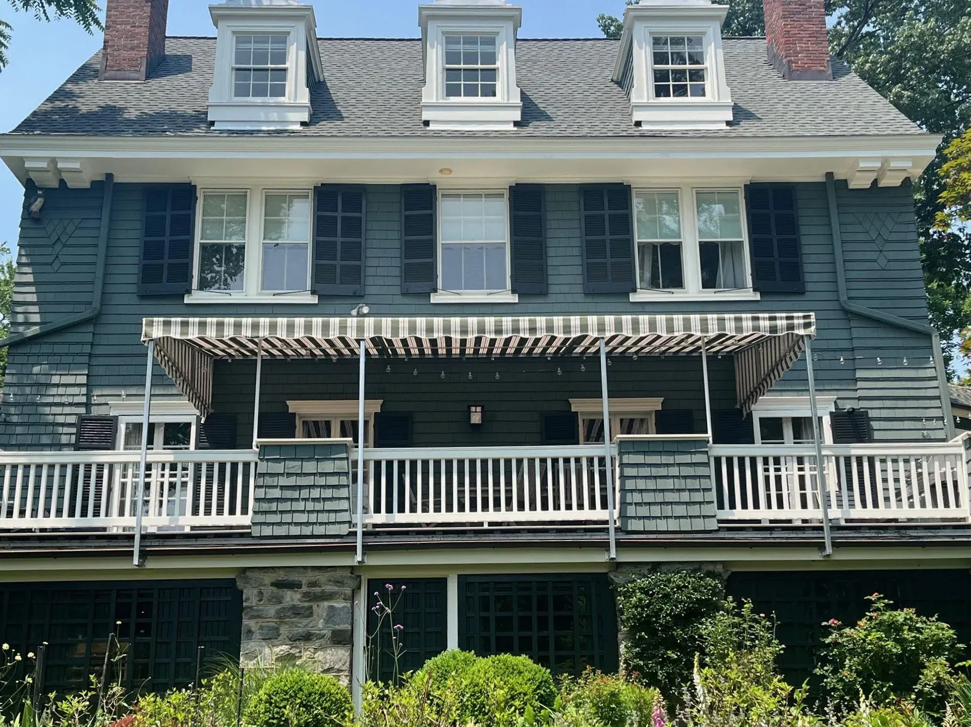 A large, dark blue three-story house with a wooden porch, striped awning, and three dormer windows under a sunny sky.