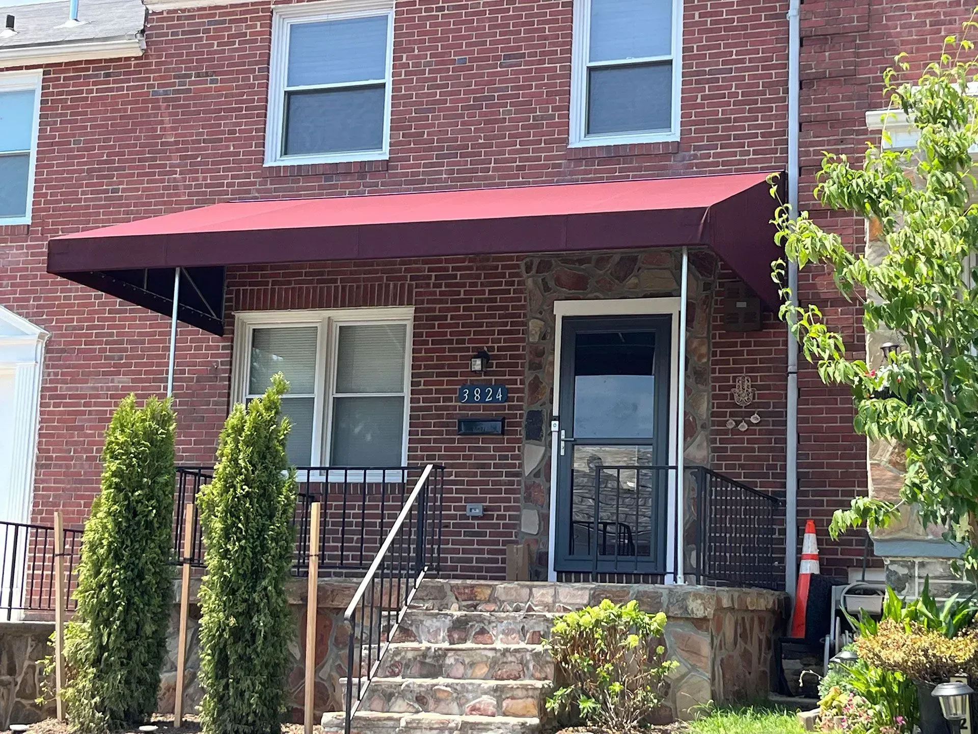 A two-story brick row house with a burgundy fabric awning, front steps, metal railing, and two small evergreen trees.